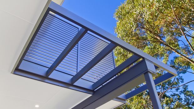 Upward-looking exterior detail of a contemporary pergola or covered outdoor structure, with dark metal framing, angled louvred roof panels, and clean white soffit surfaces against a bright blue sky and surrounding tree canopy.