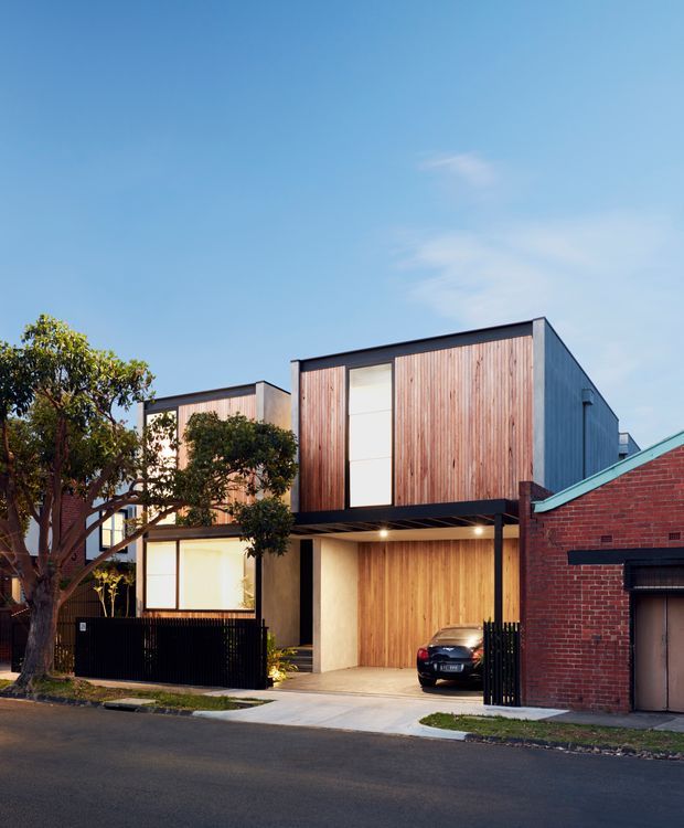 Exterior street view of a contemporary multi-level urban residence composed of box-like volumes. The facade combines vertical timber cladding, dark metal framing, large glazing, and a sheltered ground-level carport. A neighbouring red-brick building is visible at the right edge, with mature street trees and a paved driveway in front.