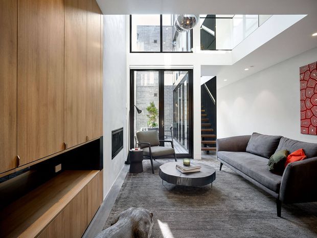 A contemporary double-height living room with a large grey sectional sofa, round coffee table, armchair, and floor-to-ceiling timber entertainment joinery. White walls, dark-framed glazing, a visible stairway and upper-level void create a bright, vertically open interior. A bold red patterned artwork adds colour contrast.