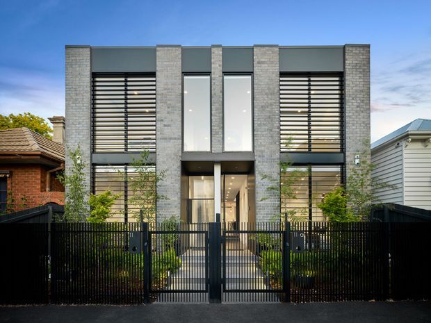 Front exterior view of a contemporary multi-storey urban residence. The symmetrical facade combines pale grey masonry piers, dark horizontal louvres, large vertical glazing, and a recessed central entry. A black metal fence and gate enclose a landscaped front path, with neighbouring brick and weatherboard homes visible on either side.