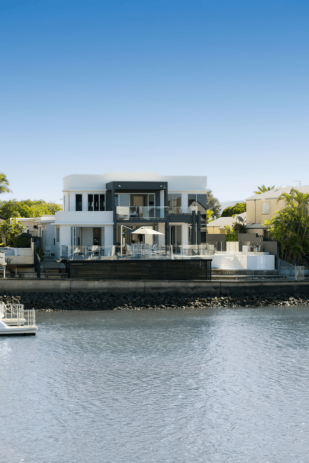 A modern multi-storey waterfront residence viewed across a calm body of water. The house has a crisp white and dark charcoal facade, extensive glazing, glass balustrades, multiple balconies and terraces, and a landscaped outdoor entertaining area with a large umbrella. A rocky seawall lines the property edge, with neighbouring homes and palms visible in the background.