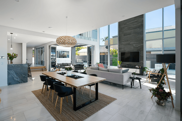 A spacious open-plan contemporary interior combining dining, living, and kitchen areas. The room features a large timber dining table with dark dining chairs, a light grey sectional sofa, pale large-format tiled flooring, a dark stone kitchen island, extensive floor-to-ceiling glazing, and a tall dark stone feature wall with a mounted television. A sculptural woven pendant light hangs over the dining table, and an upper-level void or mezzanine edge is visible.