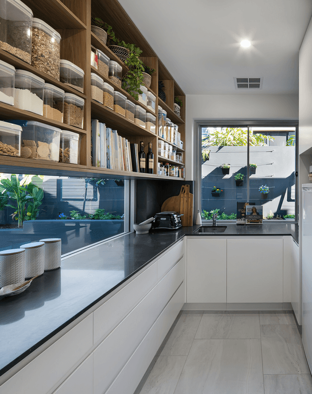 A compact contemporary kitchen with white handleless base cabinetry, dark stone-look benchtops, a stainless-steel sink, and open timber shelving filled with pantry containers, cookbooks, bottles and plants. A large window and mirrored splashback bring views of a planted courtyard into the space. Light tiled flooring and recessed ceiling lighting create a bright, practical interior.