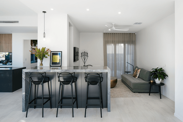 Bright open-plan contemporary interior with a marble-look kitchen island and three black bar stools in the foreground, opening to a compact living area with a grey sofa, black side table, indoor plant and sheer floor-to-ceiling curtains. White walls and ceiling, pale timber-look flooring, recessed lighting and a ceiling fan create a clean, airy composition. A partial kitchen and dining area are visible to the left.