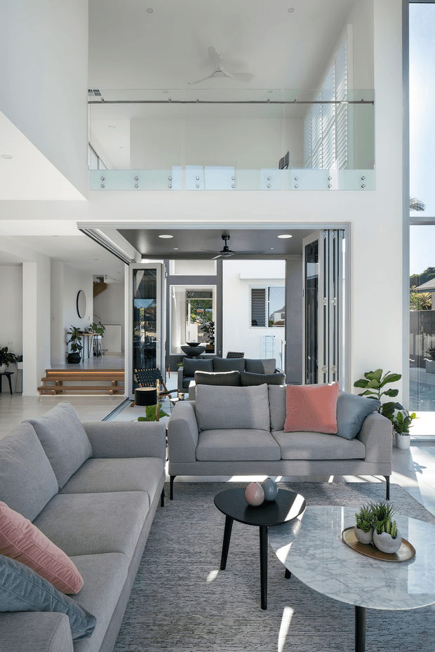 A bright, double-height contemporary living room with a large grey sectional sofa, matching two-seater sofa, black round coffee table, and marble oval coffee table. Floor-to-ceiling glazing and folding doors open toward an outdoor pool area, while a glass-balustraded mezzanine overlooks the room. White walls, pale flooring, indoor plants, and abundant natural light create an airy, refined atmosphere.