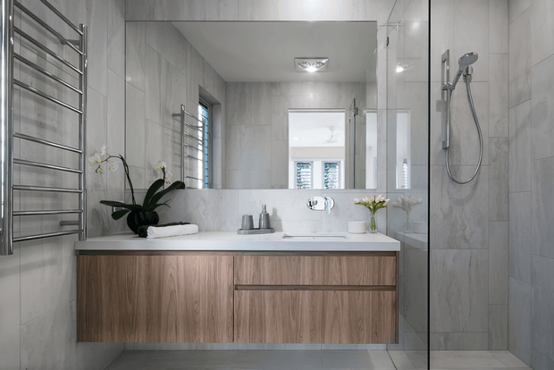 Bright contemporary bathroom featuring a wall-mounted timber vanity with a white benchtop and integrated basin, large-format light grey wall and floor tiles, a frameless glass shower enclosure with chrome fittings, a heated towel rail, and a large mirrored wall above the vanity. Natural light enters through the window, with minimal styling including orchids, towels, and small vases.