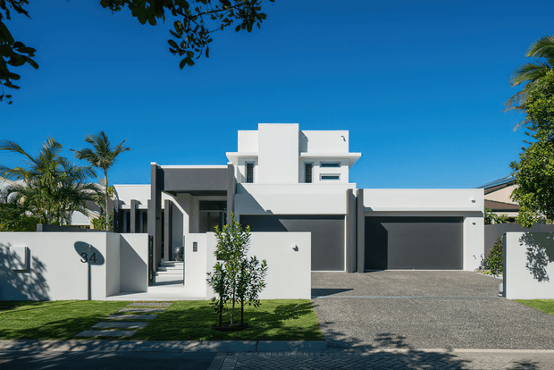 Street-facing view of a contemporary white and charcoal rendered residence with a flat roof, strong geometric massing, recessed entry, privacy walls, and an attached double garage. A paved driveway, front lawn, young tree, and tropical planting frame the minimalist facade under a clear blue sky.