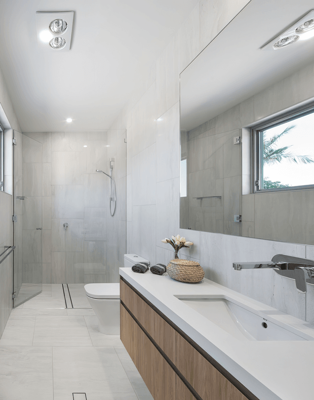 Bright contemporary bathroom with a walk-in glass shower, wall-hung toilet, large frameless mirror, and floating timber-look vanity with a white integrated benchtop and basin. Large pale wall and floor tiles create a clean, light-filled interior, with recessed and surface-mounted ceiling lighting.