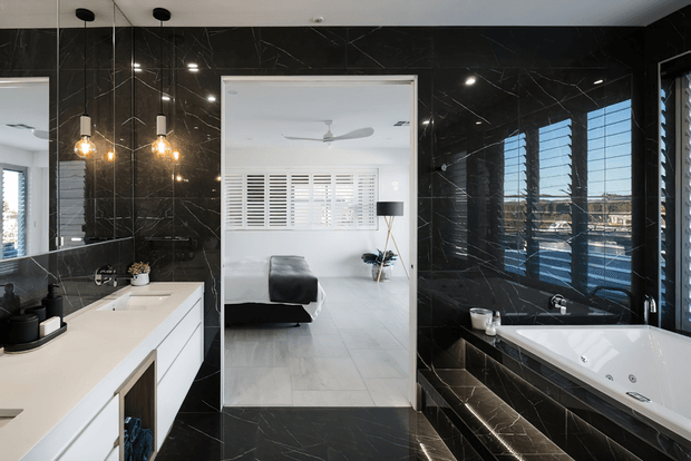 A contemporary bathroom with glossy black marble-look wall and floor surfaces, a white freestanding-style built-in spa bath, white vanity cabinetry with a pale benchtop, black fixtures, and pendant lighting. A large mirror reflects an adjacent bedroom with a bed, plantation shutters, and a ceiling fan. Dark louvre windows bring in daylight and create a dramatic, refined atmosphere.
