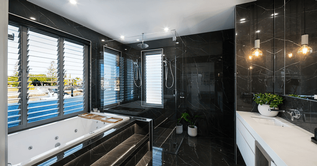 A luxurious contemporary bathroom with a large white spa bath, separate glass-enclosed shower, dark marble-look tiled walls and floors, louvred windows, and a white vanity with a dark countertop. Warm pendant lights and indoor plants add contrast to the predominantly black and white scheme.