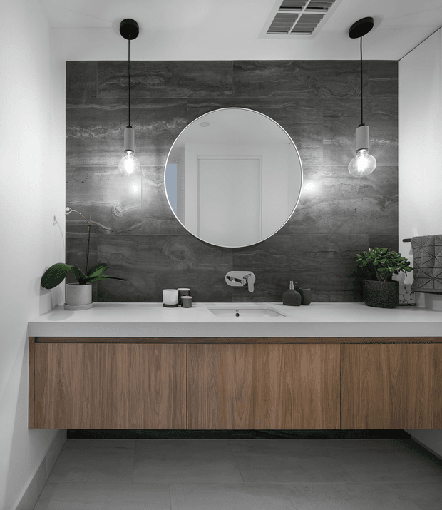 Contemporary bathroom vanity with a long floating timber-grain cabinet, white countertop and integrated basin, wall-mounted tapware, large round mirror, dark grey stone-look feature wall, pendant lights, and restrained plant styling. The space has a clean, monochromatic and refined appearance.