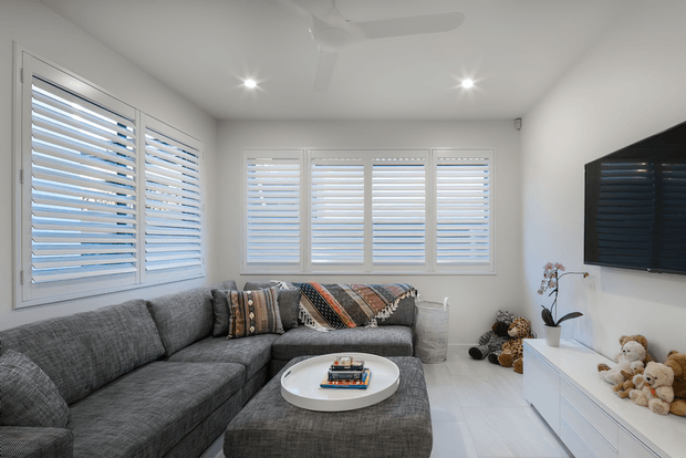 Bright, compact contemporary living room with a large grey sectional sofa and ottoman, white round coffee table, wall-mounted television, white low entertainment unit, and white plantation shutters across multiple windows. Recessed lighting and a ceiling fan create a clean, airy interior, with patterned cushions and soft toys adding informal warmth.