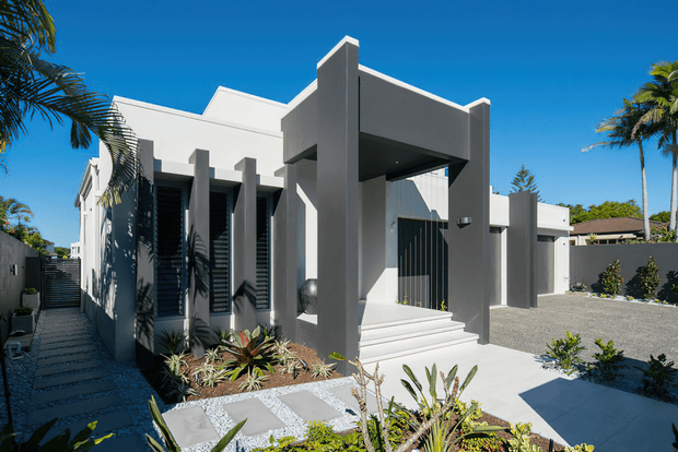 A contemporary single-storey residential facade in a tropical suburban setting, with strong geometric forms, white and grey rendered walls, deep vertical fins, a covered entry canopy, louvred window screens, broad steps, and a gravel driveway. Palm trees and drought-tolerant landscaping frame the entrance.