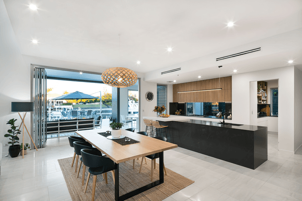 A bright, open-plan contemporary kitchen and dining area with a large black island, timber-look cabinetry, white surfaces, and a six-seat dining table. Full-height glazing opens toward an outdoor terrace and views beyond. Recessed ceiling lighting and a woven pendant provide layered illumination.