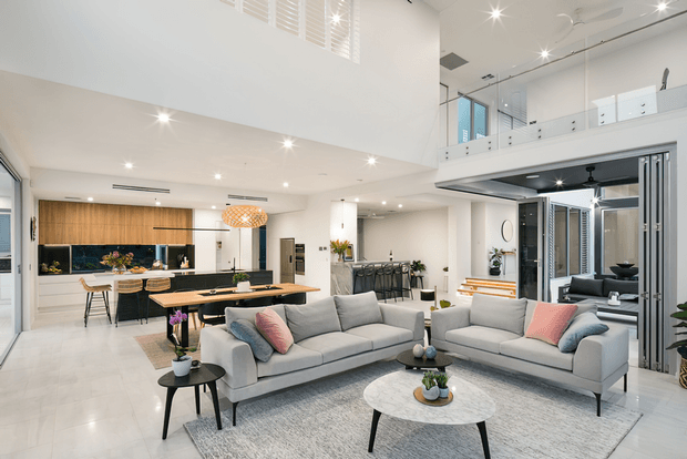 Bright open-plan contemporary living area with a large sectional-style sofa arrangement, dining table, and kitchen in the background. The room features a double-height void, mezzanine-level balustrade, extensive white finishes, pale tiled flooring, timber kitchen cabinetry, recessed lighting, and a pale stone coffee table. A partially enclosed outdoor or transitional area is visible at right.
