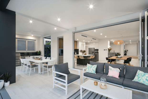 Bright open-plan contemporary living area with a grey sectional sofa, white coffee table, and white-framed armchair opening onto a dining area and modern kitchen. Large glazed doors connect the interior to an outdoor or transitional space, while white finishes, pale flooring, recessed lighting, and light timber accents create an airy coastal feel.