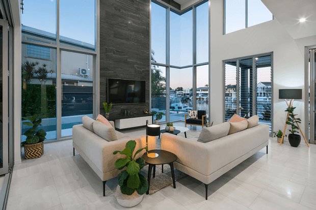 A bright, contemporary double-height living room with two cream modular sofas arranged around small round coffee tables and a television console. Floor-to-ceiling glazing wraps the corner, providing views of neighbouring buildings and water or marina-like surroundings. The room has pale tiled flooring, a dramatic dark stone-look feature wall, white walls, indoor plants, recessed lighting, and horizontal window blinds.