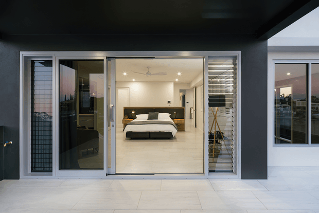 Exterior view of a contemporary residential facade with a covered tiled terrace, large sliding glass doors, and louvred screening. The glazing looks directly into a bright, minimalist bedroom with a centered bed, dark timber headboard, light tile flooring, recessed lighting, and a ceiling fan. A second glazed room is visible to the right.