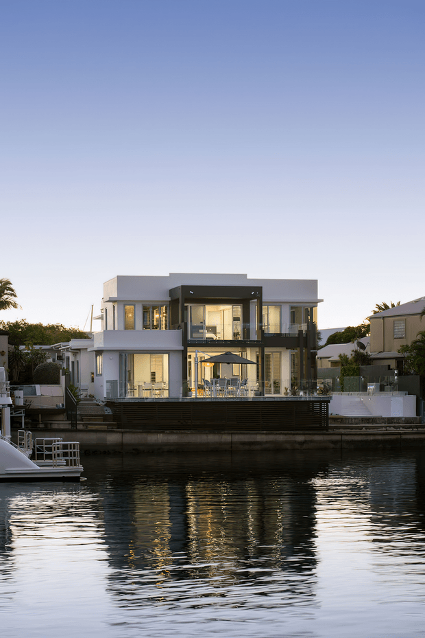 A contemporary multi-storey waterfront residence viewed across a canal at dusk. The house has a crisp white and grey facade, extensive floor-to-ceiling glazing, dark structural framing, balconies, and a large outdoor entertaining terrace with dining furniture and an umbrella. The building and its warm interior lighting are reflected in the water.