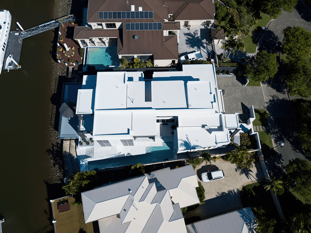 Aerial view of a contemporary waterfront residential property with expansive white flat and low-pitched roof forms, a swimming pool, landscaped outdoor areas, a private driveway, and a marina or dock along the water. Adjacent residential properties and vehicles are also visible.
