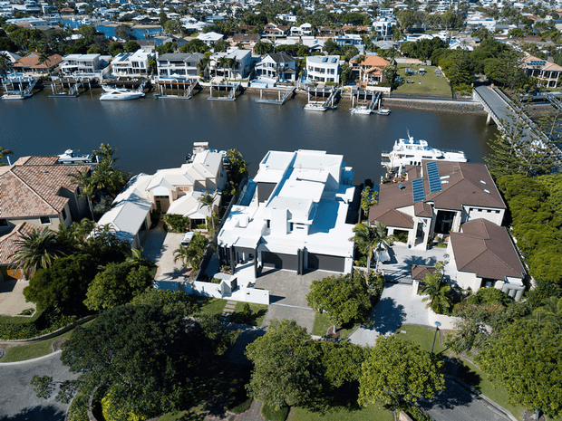 Aerial view of a suburban waterfront neighbourhood, showing several large detached homes lining a calm canal or river. The foreground includes a prominent contemporary white multi-level residence, neighbouring homes with tiled roofs, landscaped gardens, private docks, boats, and a road along the waterway.