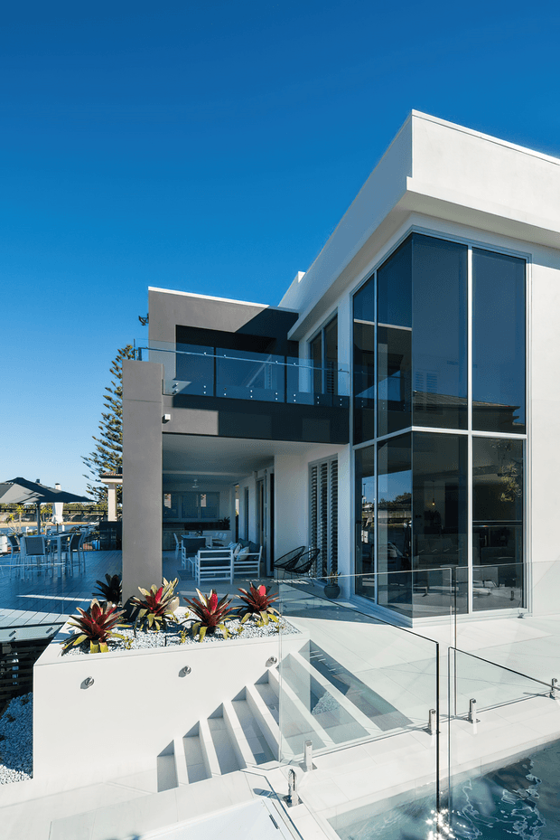 Bright exterior view of a multi-level contemporary residence with crisp white rendered walls, extensive floor-to-ceiling glazing, glass balustrades, dark metal accents, a stepped terrace, landscaped raised planter, and a swimming pool in the foreground. Outdoor dining and lounge areas are visible beside the building, with a clear blue sky and coastal-style planting.