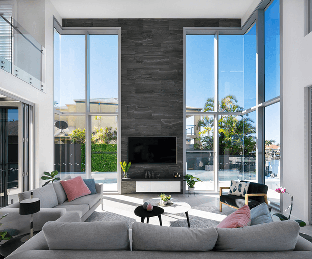 Bright contemporary living room with a large grey sectional sofa arranged around a round coffee table and wall-mounted television. Floor-to-ceiling glazing on multiple sides provides abundant daylight and views of the surrounding residential setting, while a tall dark stone feature wall forms the media backdrop. The room includes high ceilings, a partial upper-level glass balustrade, and a refined neutral palette with pink and dark blue accents.