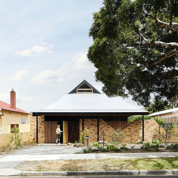 Street-facing photograph of a single-storey residential house with a broad light-coloured pitched roof, prominent triangular gable with a small clerestory window, exposed warm brick walls, dark timber screening and a covered entry/carport-like frontage. The home sits in an established suburban streetscape with a low-maintenance planted front garden, timber fencing, mature trees and neighbouring houses visible at the sides.