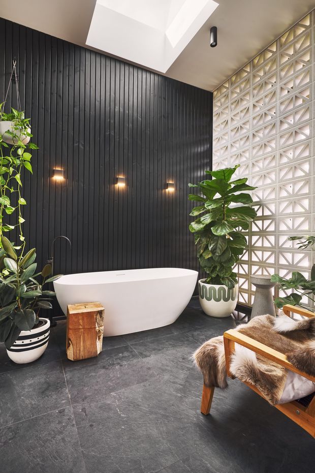 A stylish contemporary bathroom featuring a freestanding white bathtub against a black vertical timber-lined feature wall. A white geometric breeze-block wall admits filtered daylight beside the tub, while a skylight brightens the space. Dark stone-look floor tiles, warm wall sconces, potted plants, and a timber-framed fur-covered lounge chair create a dramatic yet inviting atmosphere.