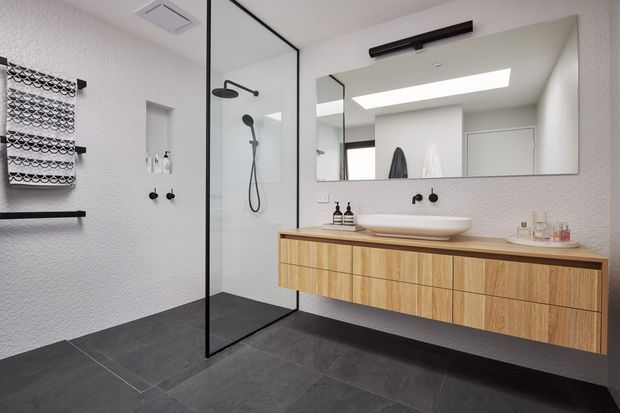 Contemporary bathroom with a floating timber vanity, white vessel basin, large frameless mirror, black tapware and lighting, walk-in glass shower, textured white walls, and large dark grey floor tiles. A skylight brings bright natural light into the room.