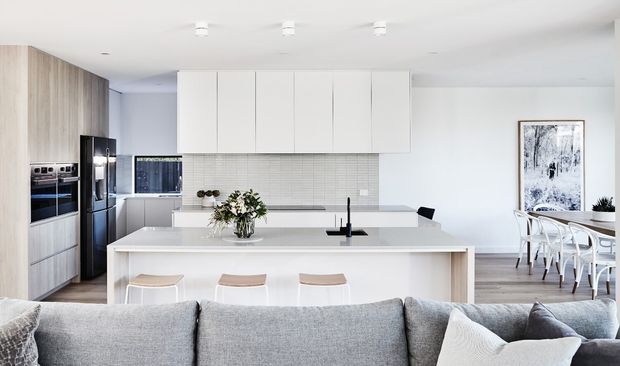 Bright open-plan contemporary kitchen viewed from the living area, featuring a large white island with seating, pale timber cabinetry, white upper cabinets, a light tiled splashback, integrated stainless-steel appliances, and a dining area beyond. A grey sofa occupies the foreground, with simple monochrome artwork and restrained decor.
