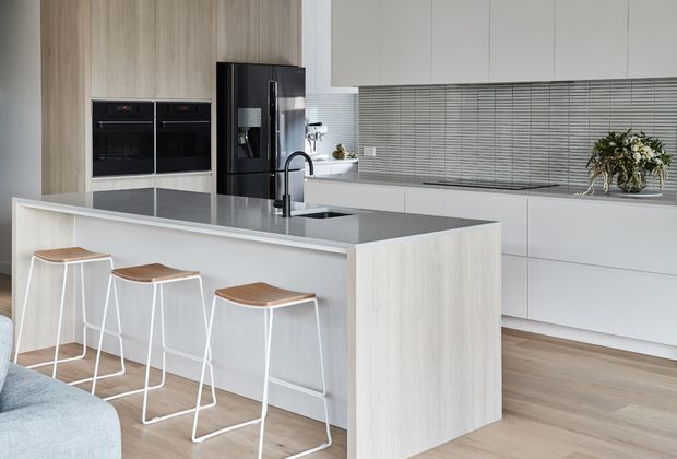 Bright contemporary kitchen with a large waterfall-edge island, integrated sink and black tap, three white-framed timber-seat stools, pale wood cabinetry, built-in black appliances, a long horizontal window with blinds, and light timber flooring. A partial sofa is visible in the foreground.