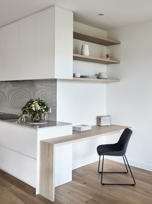 A bright, compact contemporary kitchen with white handleless cabinetry, a dark grey horizontally textured tiled splashback, and a light timber-look benchtop that extends into a built-in desk or breakfast workspace. Open timber shelves display simple ceramics, while timber flooring, a black upholstered chair, and understated decor create a clean, warm minimalist interior.