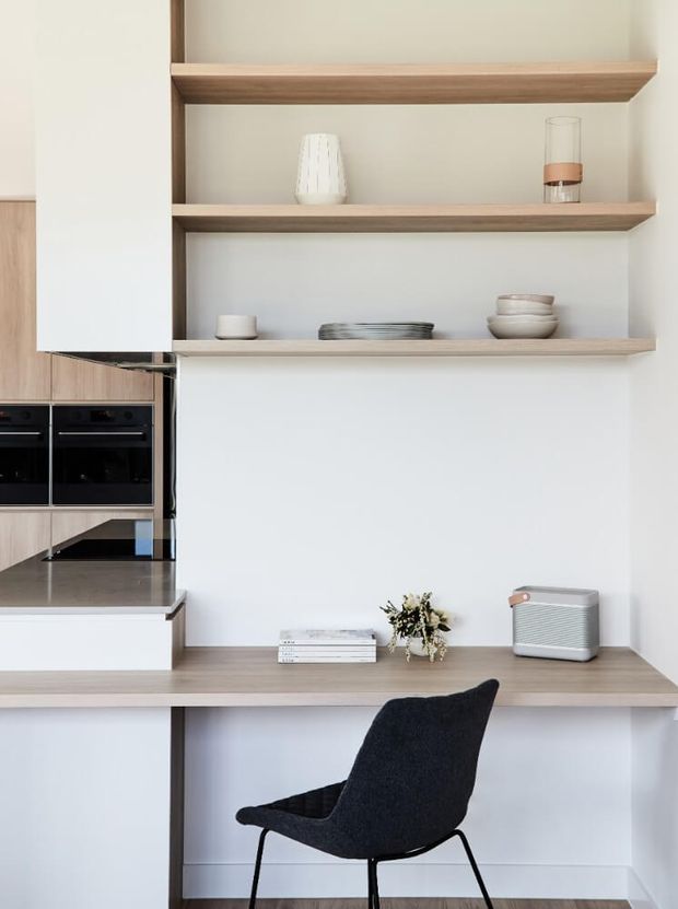A bright, minimalist kitchen and integrated study nook. White cabinetry and walls are paired with warm light timber open shelving and a long timber desk, with a black upholstered chair, built-in black ovens, and a dark stone benchtop. Open shelves display understated ceramics, glassware, books, and decorative objects.