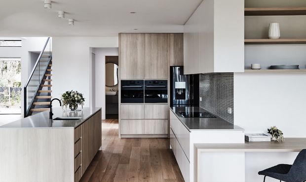 Contemporary open-plan kitchen with a large central island, integrated timber-look cabinetry, black appliances, dark mosaic-tile splashback, white benchtops, and warm timber flooring. A stairway and bright living area are visible beyond the kitchen, with open shelving and a small adjoining work or dining surface.