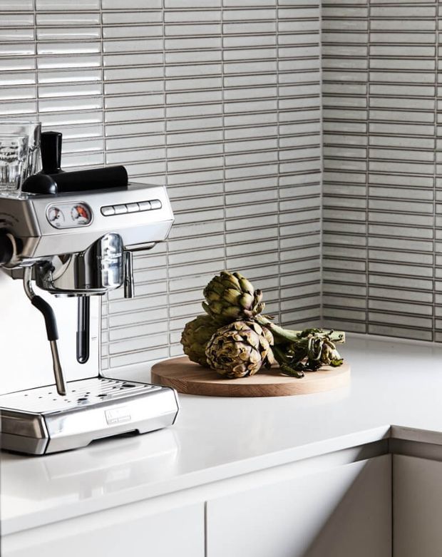 Close-up of a contemporary kitchen corner featuring a polished stainless-steel espresso machine on a white benchtop, with a round timber cutting board and artichokes. The walls are clad in narrow horizontal grey-and-white tiles, and white cabinetry with integrated handles is visible below.