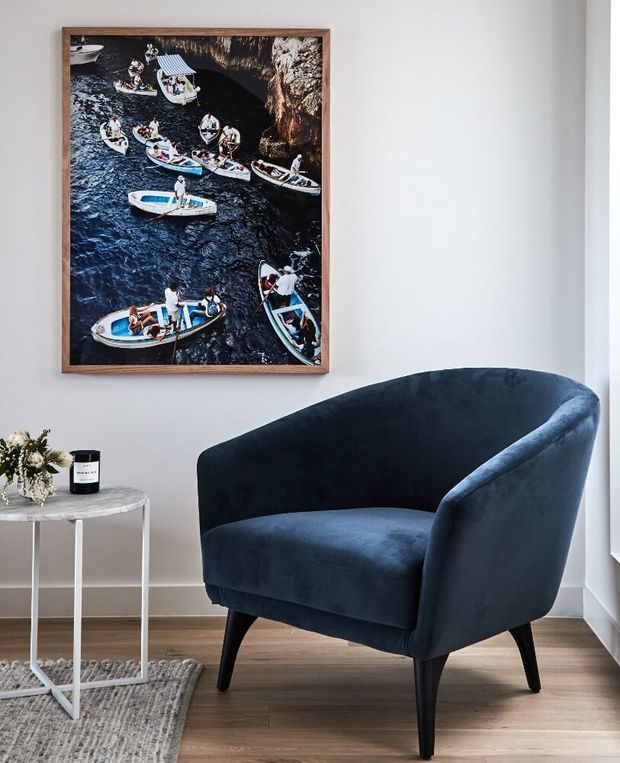 A styled interior corner featuring a deep navy-blue upholstered armchair with a curved wingback silhouette and tapered dark legs. The chair sits beside a small round white side table holding flowers and a candle, over a textured grey rug and warm timber flooring. A framed nautical photograph hangs on the light neutral wall, with bright natural light entering from the right.