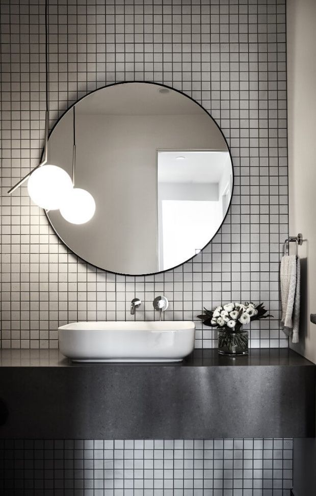 A contemporary bathroom vanity featuring a white vessel basin on a dark stone countertop, a large round mirror, black-and-white grid wall tiles, wall-mounted taps, globe pendant lighting, and a towel rail. The monochrome palette and geometric tiling create a refined, graphic look.