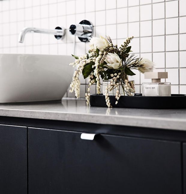 Close-up of a contemporary bathroom vanity with dark cabinetry, a pale stone countertop, a white vessel basin and wall-mounted chrome tapware. The backsplash is composed of white square tiles with dark grout. A black tray holding white floral arrangements and perfume sits on the counter.