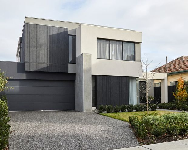 Street-facing exterior view of a contemporary two-storey residence with a flat roof, crisp rendered light-grey walls, dark vertical cladding, a dark sectional garage door, large windows, and a landscaped front garden. The home sits beside an older tiled-roof dwelling, highlighting a modern infill or extension-like urban residential context.