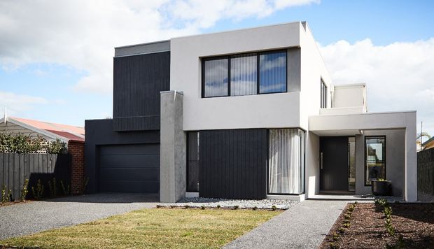 Street-facing view of a contemporary two-storey residence with a flat-roofed composition, white rendered upper volume, dark vertical cladding, large black-framed windows, an integrated garage, and a covered entry. The front landscape includes gravel paths, lawn, and simple garden beds.