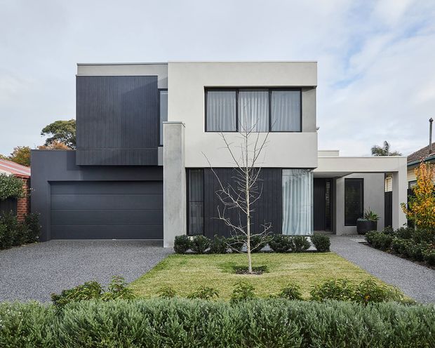 Contemporary two-storey residential facade with a minimalist rectilinear composition. The exterior combines pale rendered walls with dark vertical cladding, a dark sectional garage door, large black-framed windows, and a gravel driveway. A neat front lawn, clipped hedges, and low landscaping define the approach.