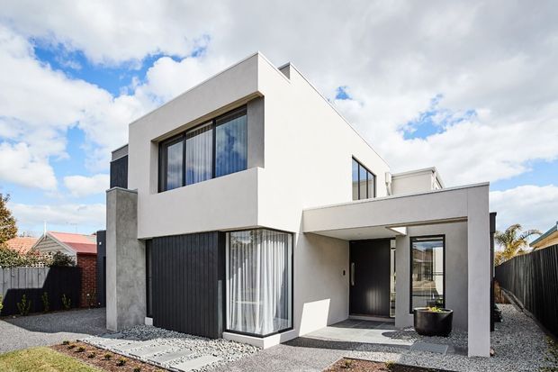 Exterior photograph of a contemporary two-storey residence with a crisp white rendered facade, charcoal vertical cladding, large glazed openings, and a projecting flat-roofed entry canopy. The front yard uses gravel and low-maintenance landscaping, with neighbouring homes visible around the property.