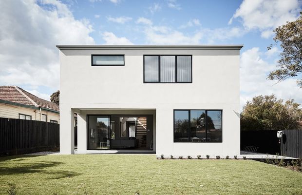 A two-storey contemporary residential rear facade with a simple white rendered rectangular form, flat roof, black-framed windows, and a recessed ground-floor outdoor living area. The house opens onto a broad lawn enclosed by dark fencing, with neighboring suburban homes visible at the sides.