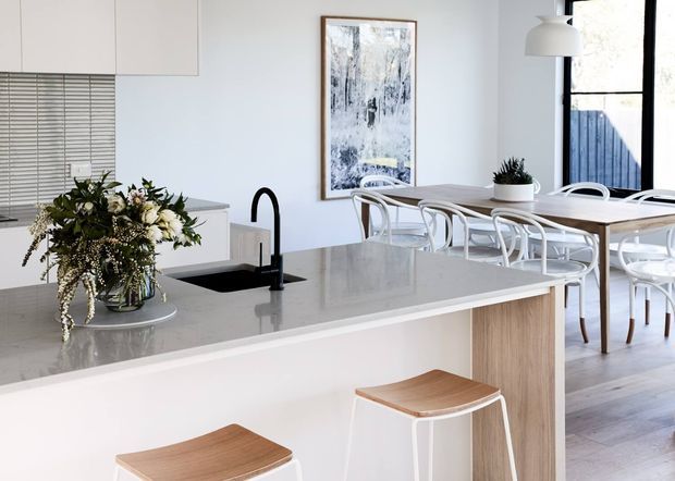 Bright, contemporary open-plan kitchen and dining area with a large pale stone island, white cabinetry, black gooseneck tap, timber-fronted island ends, and white stools with timber seats. A light timber dining table and white bentwood-style chairs sit beside large windows, complemented by an abstract artwork, pendant light, and floral arrangement.