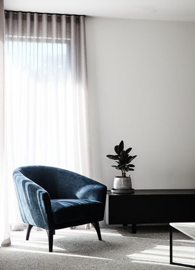 A bright, minimalist living room corner with a deep blue upholstered armchair beside a low dark built-in or freestanding media console. Floor-to-ceiling sheer curtains filter strong daylight, while a potted plant and part of a low coffee table complete the calm contemporary setting. Neutral white walls and carpet provide a simple backdrop.