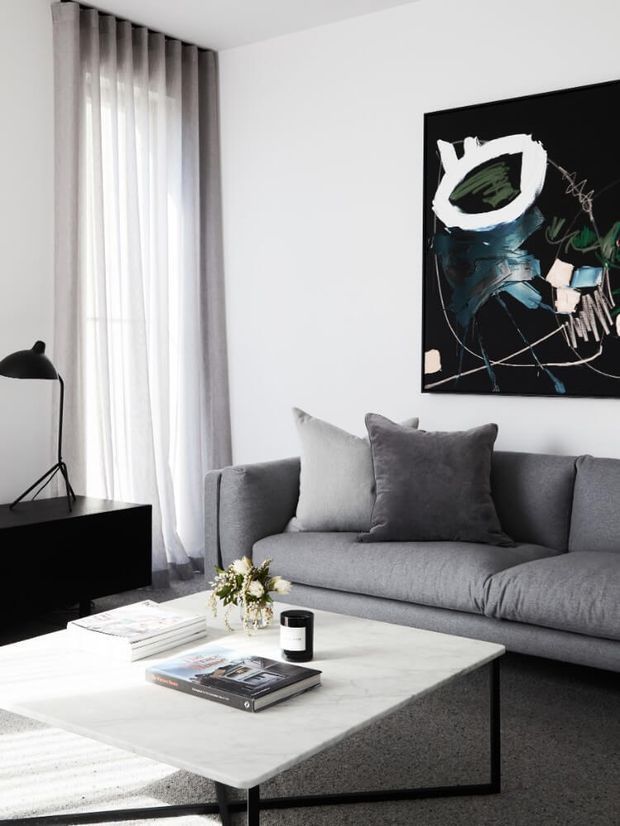 A refined contemporary living room with a grey upholstered sofa, layered grey cushions, a large pale stone coffee table, and a black low media console. Floor-to-ceiling sheer and grey drapery frame a tall window, while a bold abstract artwork and sculptural black lamp add graphic accents. The palette is predominantly white, grey, black, and light stone with a calm, polished atmosphere.