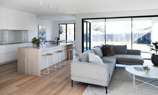 Bright open-plan contemporary living and kitchen space with pale timber flooring, white handleless cabinetry, a large island with bar seating, and a grey sectional sofa facing expansive black-framed glazing to the garden.