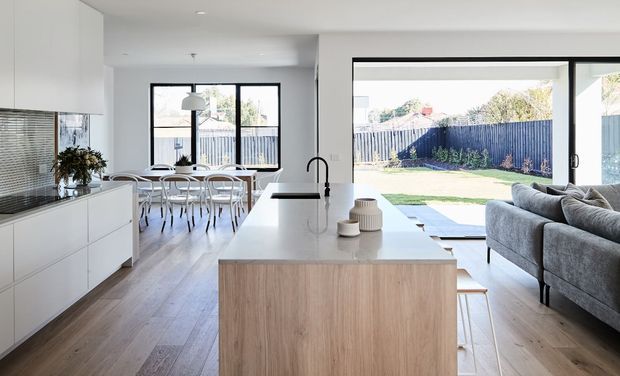 Bright open-plan contemporary interior showing a white kitchen with a large light-wood island, integrated cabinetry, pale timber flooring, and a dining area beyond. Full-height sliding glass doors open to a fenced backyard, while a grey sectional sofa occupies the living area. The space is airy and minimally styled with strong indoor-outdoor connection.
