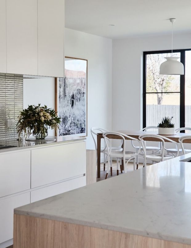 Bright, refined open-plan kitchen and dining area with white flat-panel cabinetry, a pale stone island benchtop, light timber island joinery, and a timber dining table surrounded by white chairs. Large black-framed windows, a sculptural pendant light, mirrored splashback, artwork, and a floral arrangement add contemporary character.
